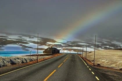 Une voiture rouge roule sur une route côtière offrant une vue spectaculaire sur les fjords norvégiens, sous un ciel bleu clair parsemé de quelques nuages. Les montagnes majestueuses se dressent en arrière-plan, invitant à l'aventure et à la découverte des paysages époustouflants de la Norvège.