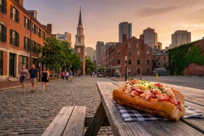 Un lobster roll sur une table en bois avec une rue pavée historique de Boston et l'horizon au coucher du soleil.