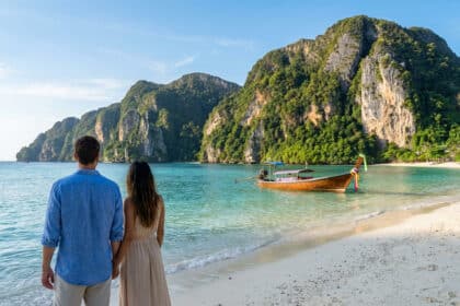 Un couple de dos sur une plage de sable blanc face à une baie turquoise avec un longtail boat et des falaises verdoyantes à Ko Phi Phi.