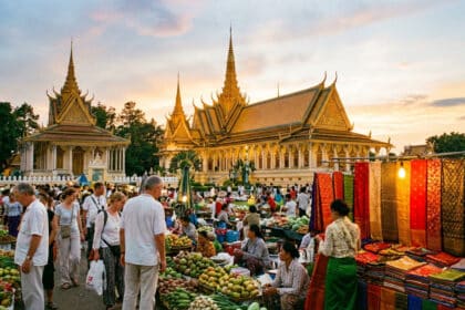 Un marché de rue animé à Phnom Penh au Cambodge, avec des habitants et des touristes, vendant des fruits et textiles devant le Palais Royal sous un ciel de coucher de soleil.