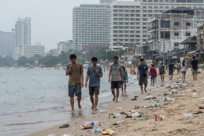 Plage sablonneuse jonchée de déchets plastiques, où des gens marchent, avec des immeubles modernes et délabrés en arrière-plan sous un ciel gris.