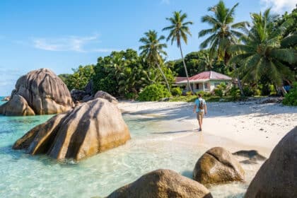 Homme avec sac à dos marchant sur une plage des Seychelles, sable blanc, eau turquoise, rochers granitiques et palmiers.