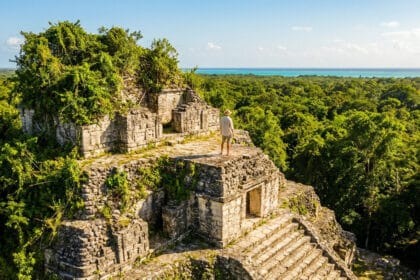 Un homme sur une ruine maya envahie par la jungle, regardant la forêt dense et la mer turquoise sous un ciel bleu.