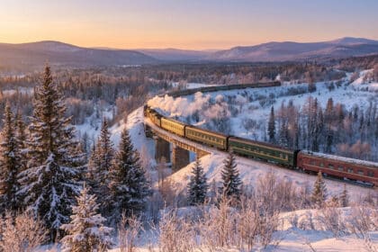 Long train vert et rouge sur un viaduc enneigé au milieu d'une vaste forêt de conifères, éclairé par un soleil de crépuscule en Sibérie.