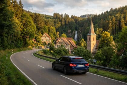 Voiture bleue sur route sinueuse en Forêt-Noire. Village, église et cascade majestueuse entourés de forêts verdoyantes sous lumière dorée.