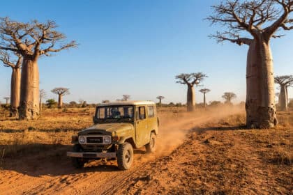 Un 4x4 Toyota beige roule sur une piste de terre rouge parmi de grands baobabs sous un ciel bleu clair à Madagascar.