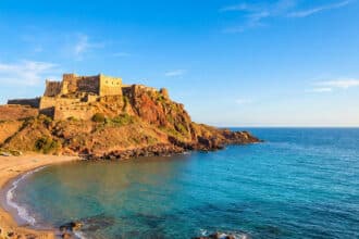 Château médiéval de Castelsardo sur une falaise rocheuse surplombant une plage de sable et une mer turquoise sous un ciel bleu.
