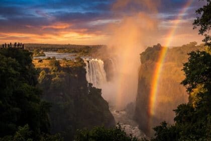 Vue panoramique des Chutes Victoria au coucher du soleil, avec un arc-en-ciel, un éléphant et des visiteurs sur une plateforme.