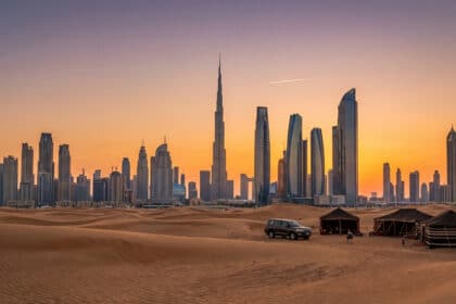 Vue panoramique de Dubaï au coucher du soleil depuis le désert, avec des dunes, un 4x4, des tentes et la skyline incluant Burj Khalifa.