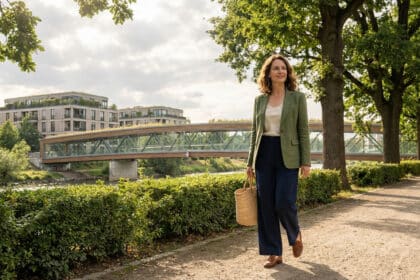 Femme élégante et souriante marchant dans un parc urbain, bâtiments modernes à toits verts et pont en arrière-plan.