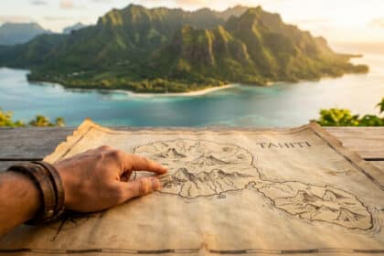 Main pointant une carte vintage de Tahiti sur une table en bois, avec des montagnes verdoyantes et un lagon turquoise en arrière-plan.