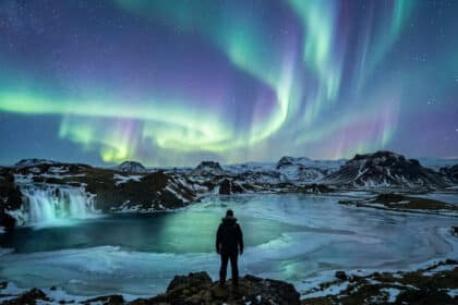 Une personne regarde les aurores boréales au-dessus d'une cascade gelée et d'un paysage montagneux en Islande.