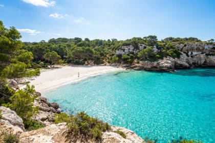 Vue panoramique sur la plage de Cala Turqueta avec son eau turquoise cristalline, entourée de falaises et de pins.