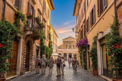 Touristes marchant dans une rue pavée fleurie menant à la basilique Santa Maria in Trastevere au crépuscule.