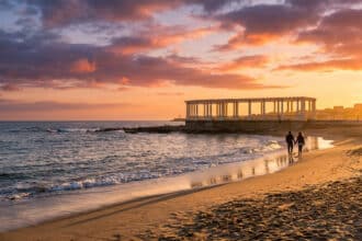 Un couple marche main dans la main sur une plage de Porto au coucher du soleil devant une pergola historique.