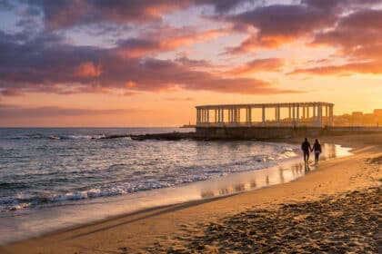 Un couple marche main dans la main sur une plage de Porto au coucher du soleil devant une pergola historique.