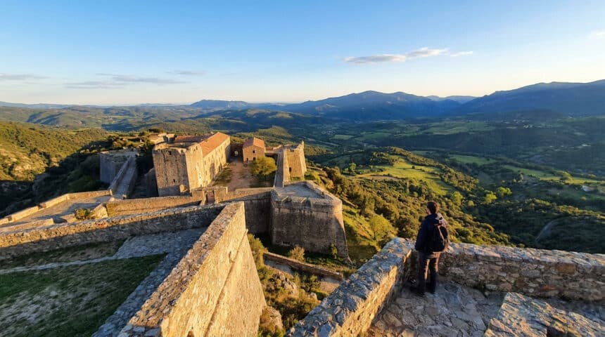 Un randonneur admire le Fort de Bellegarde sous une lumière dorée, surplombant les montagnes des Pyrénées.