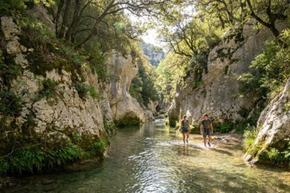 Deux randonneurs marchant dans les eaux peu profondes des Gorges du Toulourenc bordées de hautes falaises rocheuses.