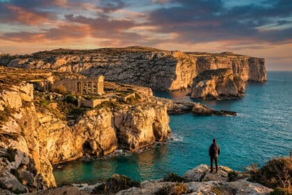 Un homme debout sur une falaise rocheuse face à une mer turquoise et des ruines antiques au coucher du soleil à Gozo.