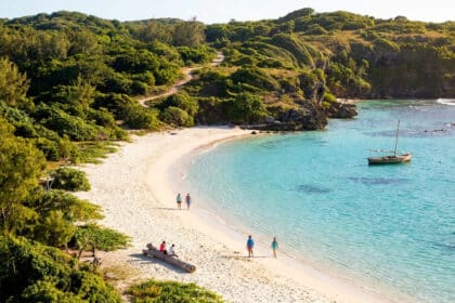 Vue plongeante d'une plage de sable blanc bordée de collines verdoyantes et d'une mer turquoise avec un petit bateau.