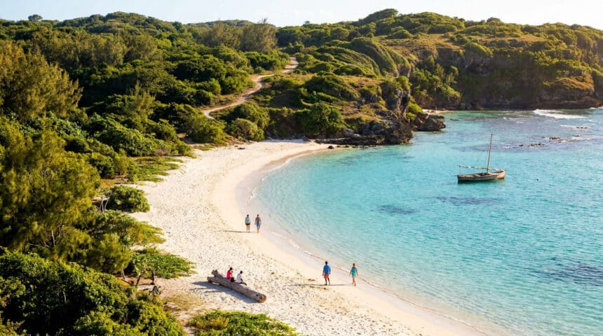Vue plongeante d'une plage de sable blanc bordée de collines verdoyantes et d'une mer turquoise avec un petit bateau.
