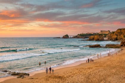 Vue panoramique d'une plage à Biarritz au coucher du soleil avec des personnes se promenant et des surfeurs dans l'eau.