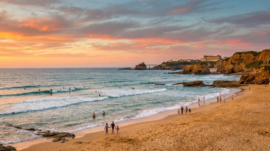 Vue panoramique d'une plage à Biarritz au coucher du soleil avec des personnes se promenant et des surfeurs dans l'eau.