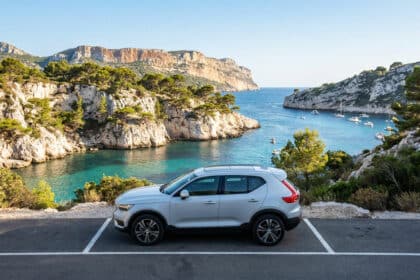 Une voiture garée sur un parking surplombant la calanque de Port-Miou avec des bateaux dans l'eau turquoise en arrière-plan.